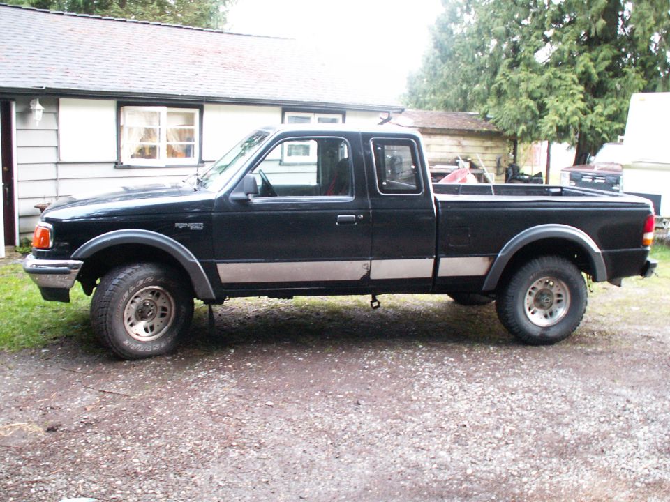Black 1990s Ford Ranger pickup truck, side view, parked on gravel in front of a house