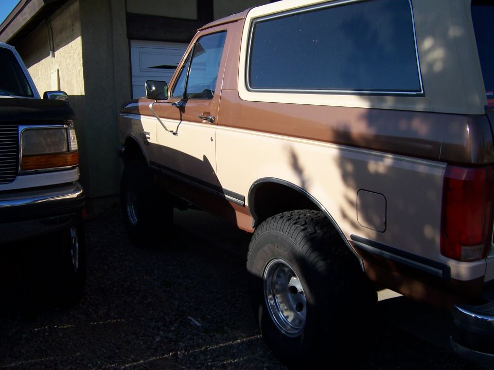 Two-tone tan and brown lifted Ford Bronco SUV parked on gravel beside a house, rear three-quarter view