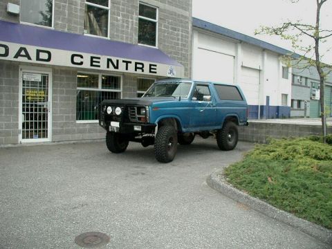 1985 Ford Bronco - A modified blue Ford Bronco is parked outside a commercial building with a purple awning.