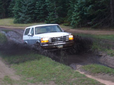 1995 Ford Bronco - A white 1995 Ford Bronco navigates a muddy trail, kicking up a large spray of water and dirt.