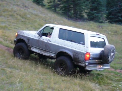 1995 Ford Bronco - A white Ford Bronco is seen driving off-road on a grassy incline, covered in mud.