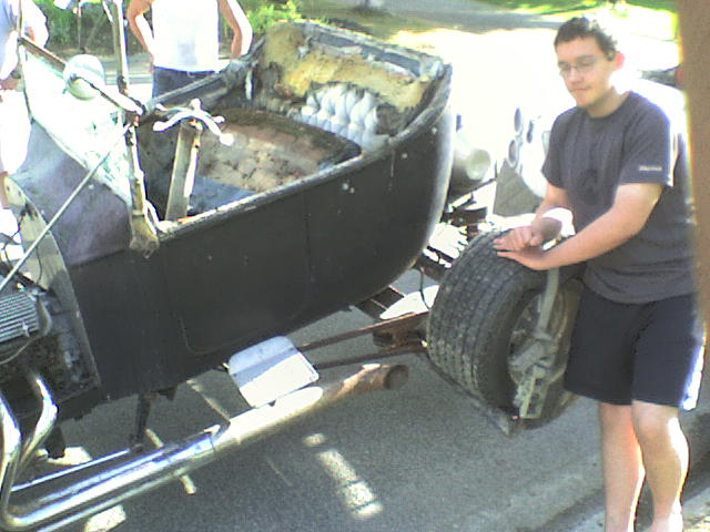 Black vintage car body, partially disassembled, with a person touching the front tire. The interior is exposed and worn