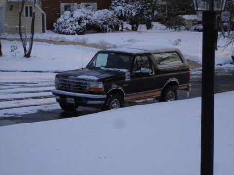 1995 Ford Bronco - Black 1995 Ford Bronco sits parked on a snow-covered road with houses and trees in the background.