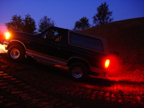 1995 Ford Bronco - A dark-colored Ford Bronco is shown driving on a dirt path as dusk settles, with its lights illuminating the surroundings.