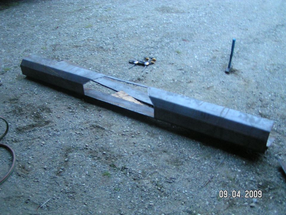 Bare steel rear bumper cut and sectioned in the middle, lying on gravel with tools nearby