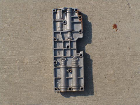 1996 Ford Bronco - A metal transmission valve body part is shown lying on a concrete surface, casting a shadow.