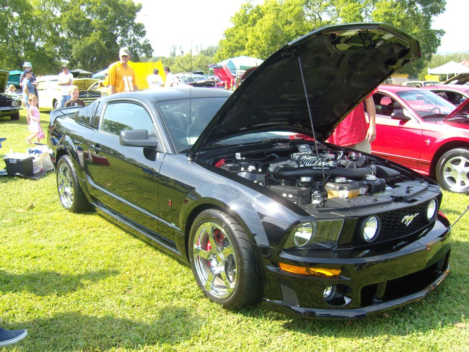 Black Ford Mustang with hood open, showing engine and chrome wheels, parked on grass at an outdoor event