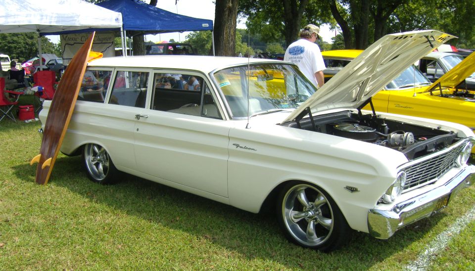 White station wagon with a surfboard leaning on its side, chrome wheels, hood open, parked on grass