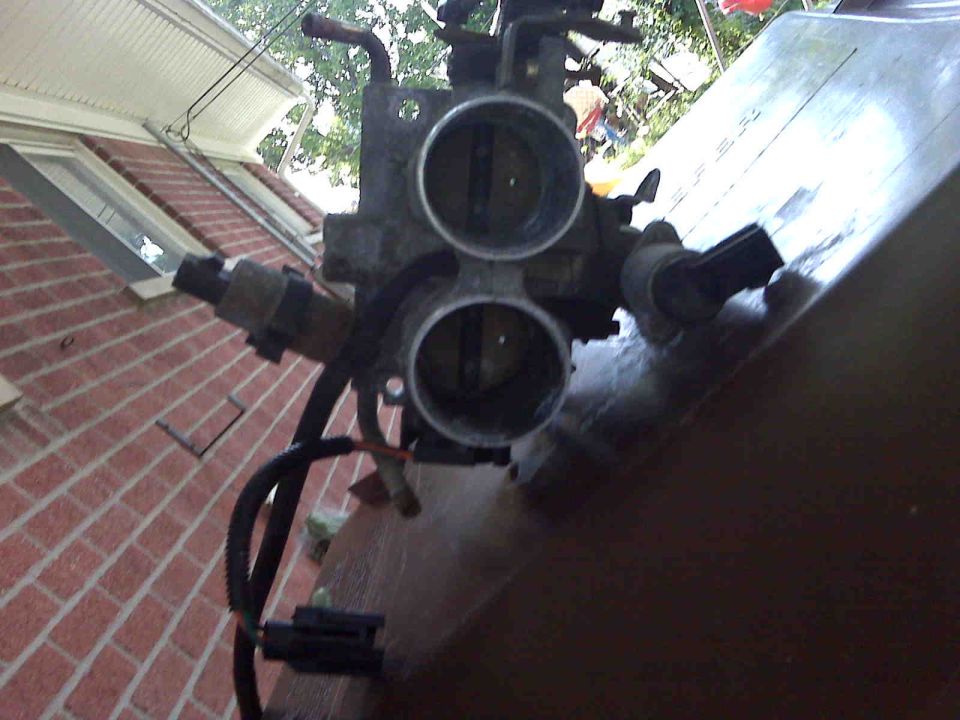 Close-up of a dual throttle body with hoses and electrical connectors attached, set against a brick wall and some greenery