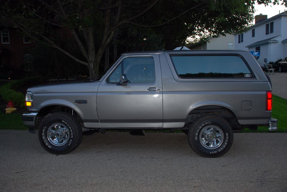 Gray Ford Bronco parked on asphalt, side view. Visible "Bronco" badge on fender and "Ford" oval on rear quarter panel