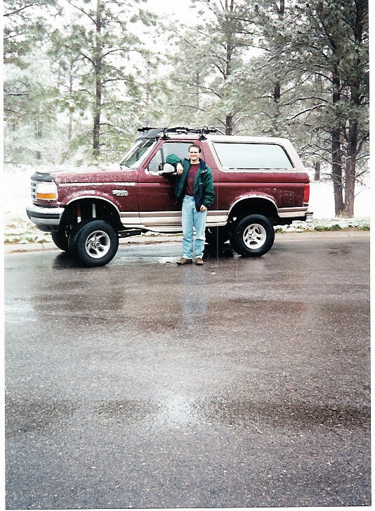 Man in denim jacket stands beside a lifted maroon Ford Bronco on a wet snowy lot