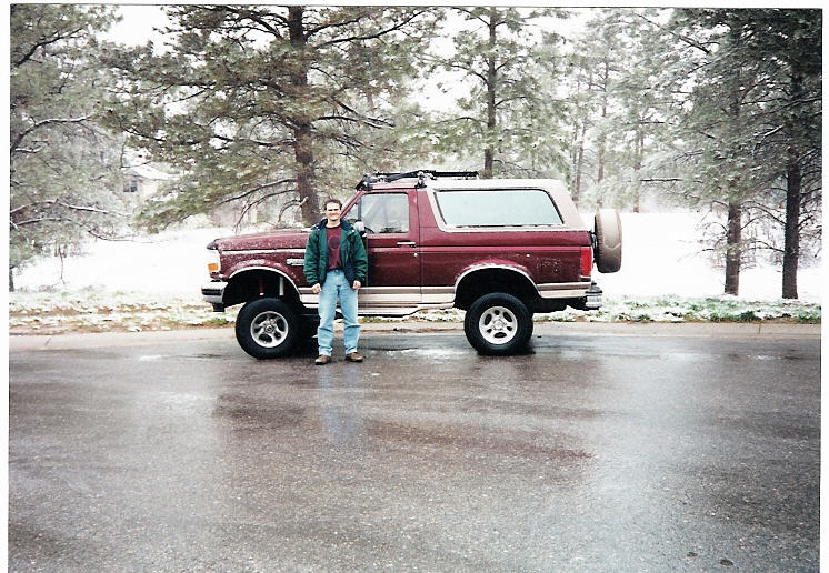 Man standing in front of a lifted maroon Ford Bronco, full side view on wet snowy pavement