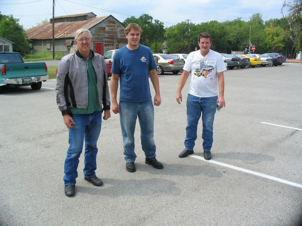Three men standing in a parking lot with cars and an old building behind them