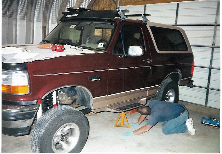 Maroon Ford Bronco SUV on jack stands in garage with person working on driver's side