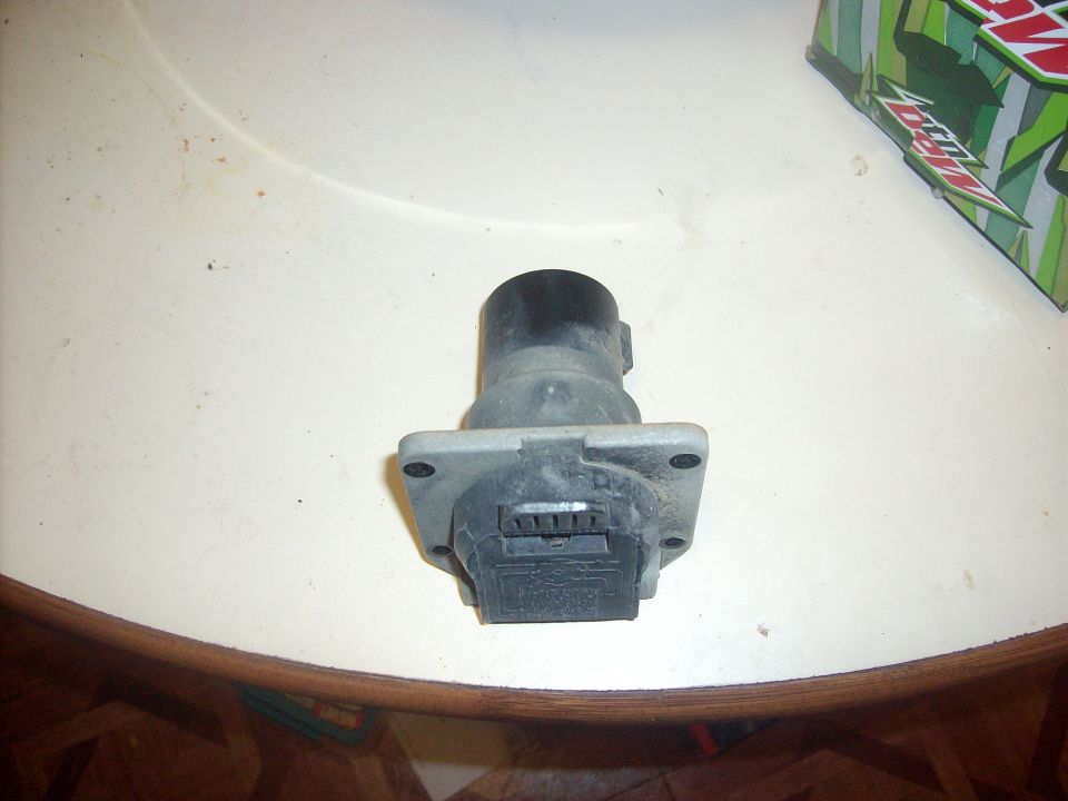 Trailer hitch connector with a black top and gray base, sitting on a table. A green and white box is in the background