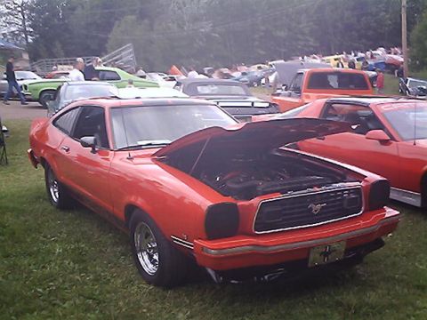 1978 Ford Mustang II - A red coupe with its hood open is parked on grass, with other vehicles and people in the background.