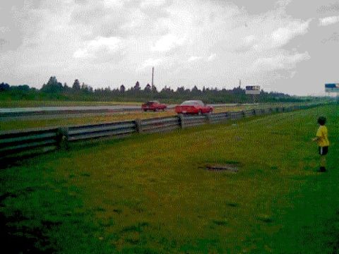 1978 Ford Mustang II - Two red cars are on a race track with a young spectator standing on the grassy shoulder.