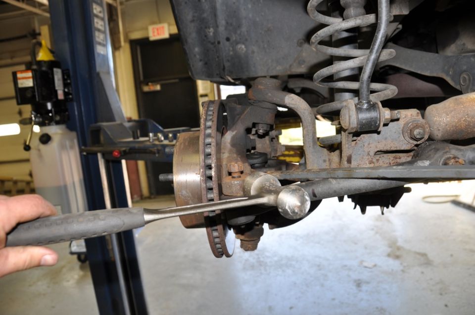 Close-up of a vehicle's front suspension and brake rotor, with a hand holding a tool near the ball joint