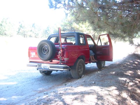 1988 Ford Bronco - Three-quarter rear view of a red Ford Bronco with its hardtop removed and roll bar exposed, parked on a dirt turnout under pine trees with…