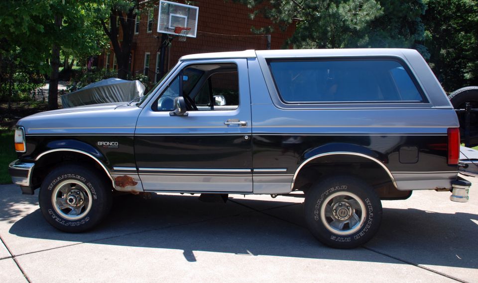 Side view of a two-tone silver and black 1995 Ford Bronco parked on a driveway. Some rust is visible on the lower body panel