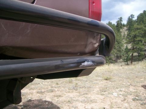 1993 Ford Bronco - The rear of a vehicle is shown with a metal bumper and a red tail light visible against a backdrop of trees and sky.