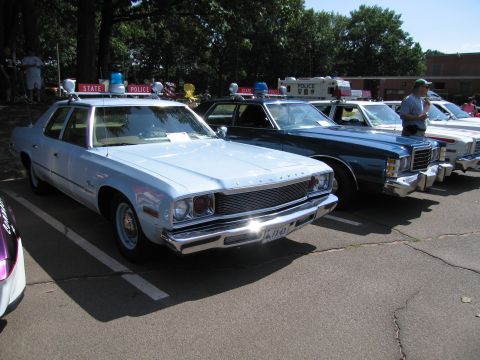 1991 Ford LTD Crown Victoria - light-blue Plymouth State Police sedan parked beside other classic police cruisers under trees at a car show.