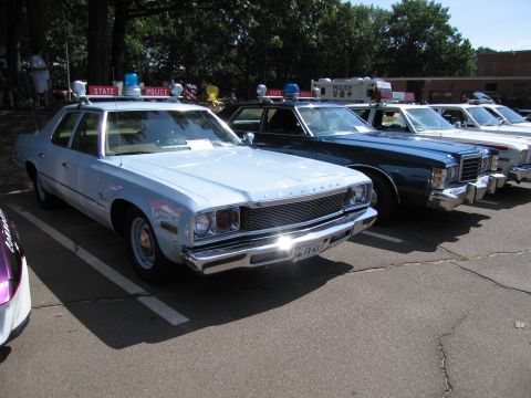 1991 Ford LTD Crown Victoria - light-blue Plymouth State Police sedan with roof light leads a row of vintage police cars at a car show.