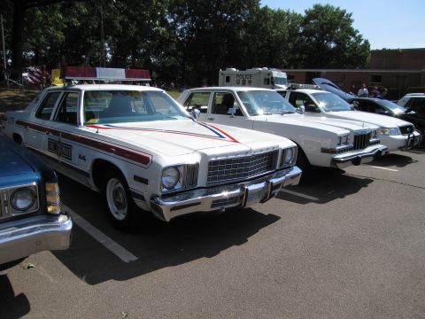 1991 Ford LTD Crown Victoria - white Plymouth Fury sedan with red and blue stripes and a roof lightbar, parked among other cars at a show.