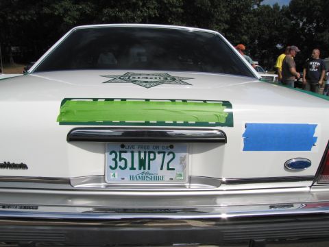 1991 Ford LTD Crown Victoria - Rear deck of a white sedan showing a sheriff star decal and a New Hampshire 351WP72 plate, taped trim, people behind.