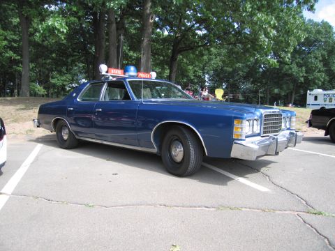 1991 Ford LTD Crown Victoria - dark blue 1970s Ford full-size sedan marked as a State Police car, with red roof signs and spotlights, parked on asphalt under trees.