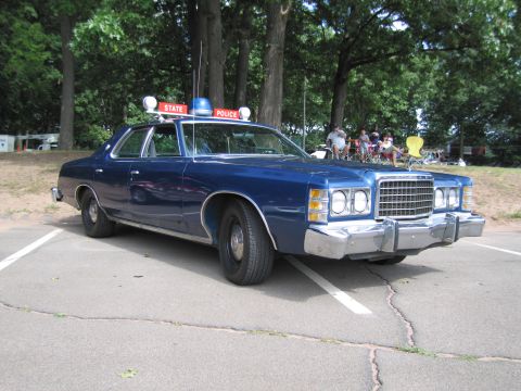 1991 Ford LTD Crown Victoria - Front three-quarter view of a blue 1970s Ford State Police sedan showing dual stacked headlights, chrome bumper guards and STATE POLICE roo…