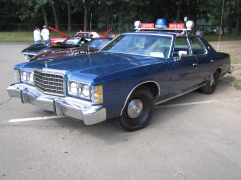 1991 Ford LTD Crown Victoria - Front three-quarter view of a blue 1970s Ford State Police sedan at a car show, with other classic cars and people in the background.