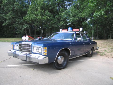 1991 Ford LTD Crown Victoria - blue 1970s Ford State Police sedan photographed from the front three-quarter angle in a paved lot with trees behind.