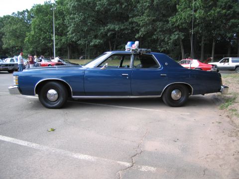 1991 Ford LTD Crown Victoria - Full passenger-side profile of a blue 1970s Ford four-door State Police sedan with roof beacon, parked on asphalt by trees.