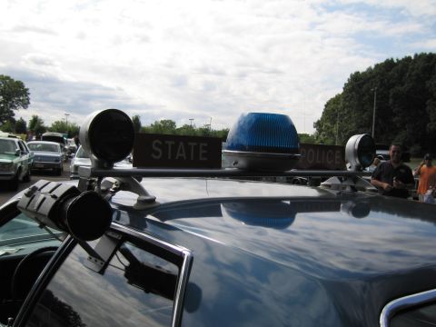 1991 Ford LTD Crown Victoria - Close-up of a police car roof showing STATE POLICE signs, a blue dome beacon and chrome spotlights against a cloudy sky.