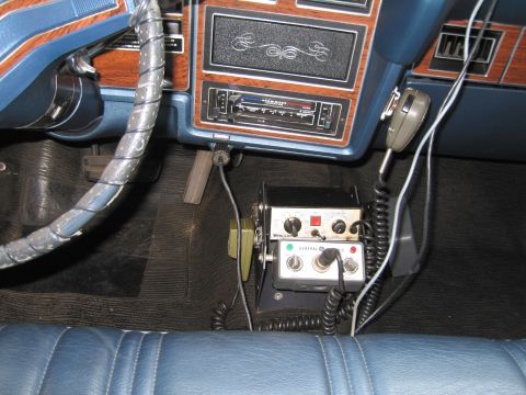 1991 Ford LTD Crown Victoria - Interior detail of a vintage car showing a floor-mounted two-way police radio with handset, beside a blue dash and woodgrain trim.