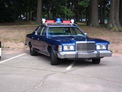 1991 Ford LTD Crown Victoria - Front three-quarter view of a blue 1970s Ford State Police sedan with quad headlights and roof beacon, parked in a tree-lined lot.