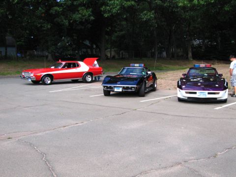 1991 Ford LTD Crown Victoria - red 1970s Ford Gran Torino with white stripe parked beside two Chevrolet Corvettes, one black and one purple-and-white, in a paved lot.