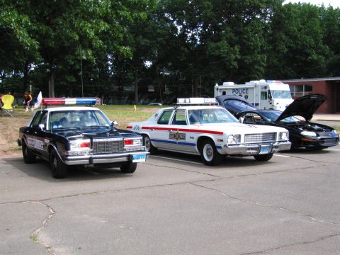 1991 Ford LTD Crown Victoria - lineup of vintage police vehicles including a black sedan, a white-and-blue patrol car, a police van and a black coupe with open hood.