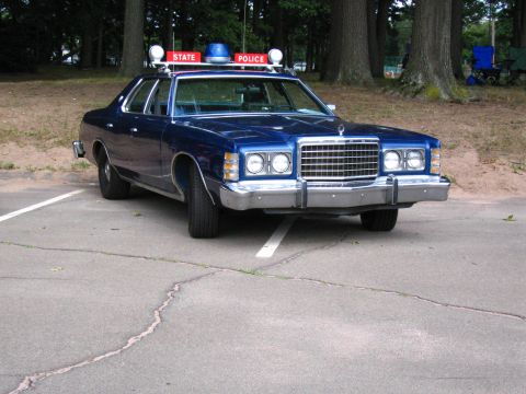 1991 Ford LTD Crown Victoria - Front three-quarter view of a blue 1970s Ford State Police sedan with quad headlights and STATE POLICE roof signs, parked by trees.