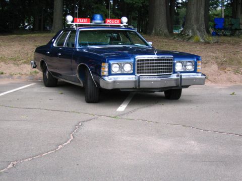 1991 Ford LTD Crown Victoria - Front three-quarter view of a blue 1970s Ford State Police sedan with chrome grille and roof beacon, parked on cracked asphalt.
