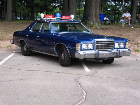 1991 Ford LTD Crown Victoria - Blue 1970s Ford full-size four-door sedan in state police livery, shown from a passenger-side front three-quarter angle in a tree-lined par…