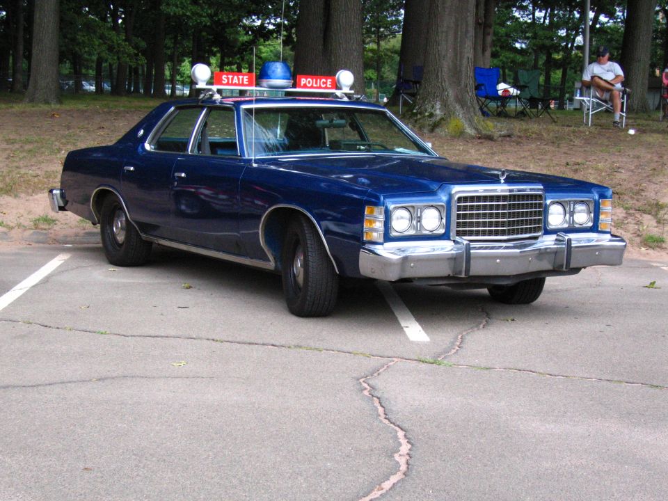 Blue 1970s Ford state police sedan with lightbar parked in a tree-lined lot