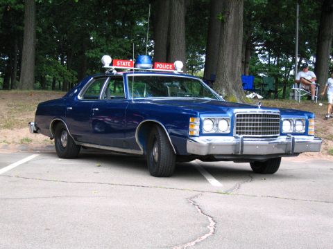 1991 Ford LTD Crown Victoria - Blue 1970s Ford LTD-style four-door police sedan parked at an angle on cracked asphalt under oak trees, viewed from the passenger front thr…