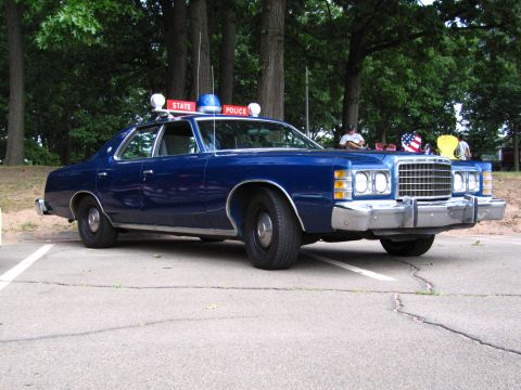 1991 Ford LTD Crown Victoria - Side-front view of a blue 1970s Ford State Police sedan parked at a show, with spectators and an American flag in the background.