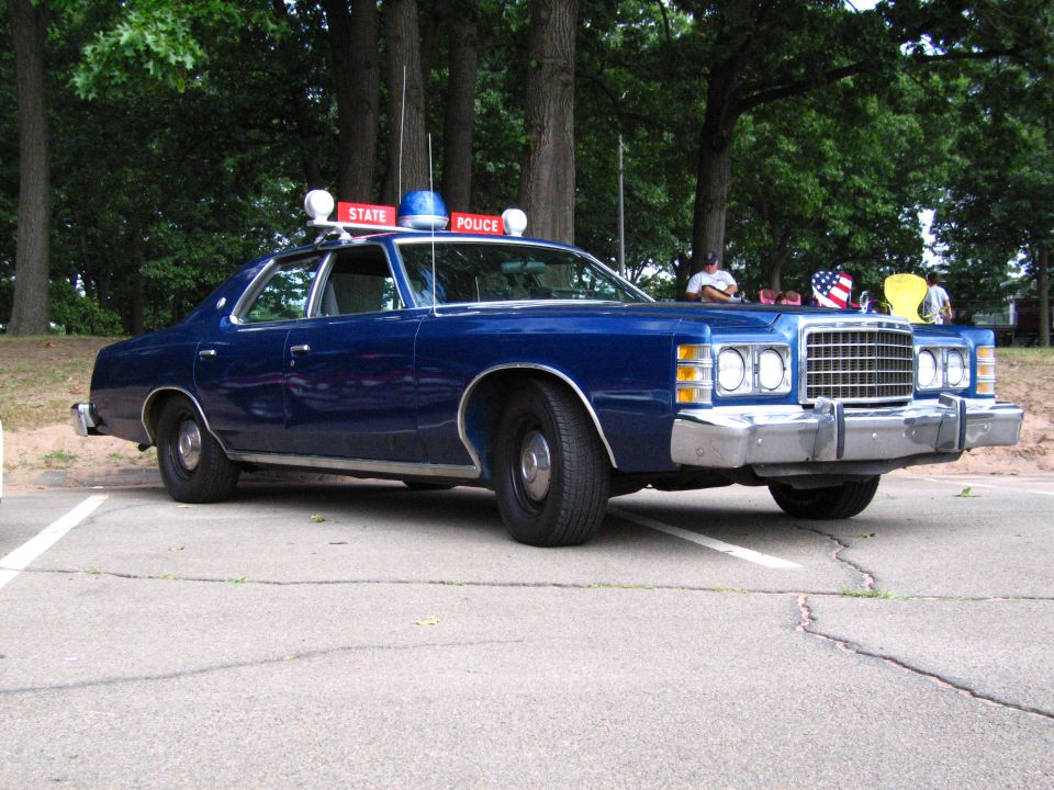 Blue Ford State Police sedan side-front view with onlookers behind