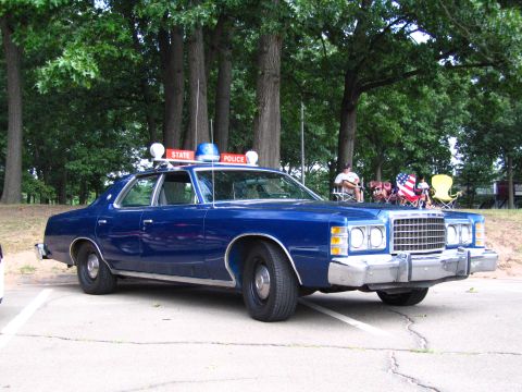 1991 Ford LTD Crown Victoria - Front three-quarter view of a blue 1970s Ford State Police sedan parked under tall trees at an outdoor car show with spectators behind.