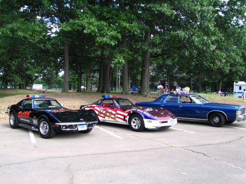 1991 Ford LTD Crown Victoria - Three show cars in a row: a black D.A.R.E. Chevrolet Corvette, a flag-graphics Corvette, and a blue 1970s Ford sedan, parked under trees.
