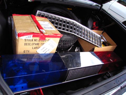 1991 Ford LTD Crown Victoria - open car trunk holding a boxed spare tire, a chrome grille, and a red-and-blue police light bar.