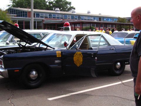 1991 Ford LTD Crown Victoria - dark blue Dodge sedan with its hood up, a yellow door shield emblem and roof beacon, with onlookers nearby.